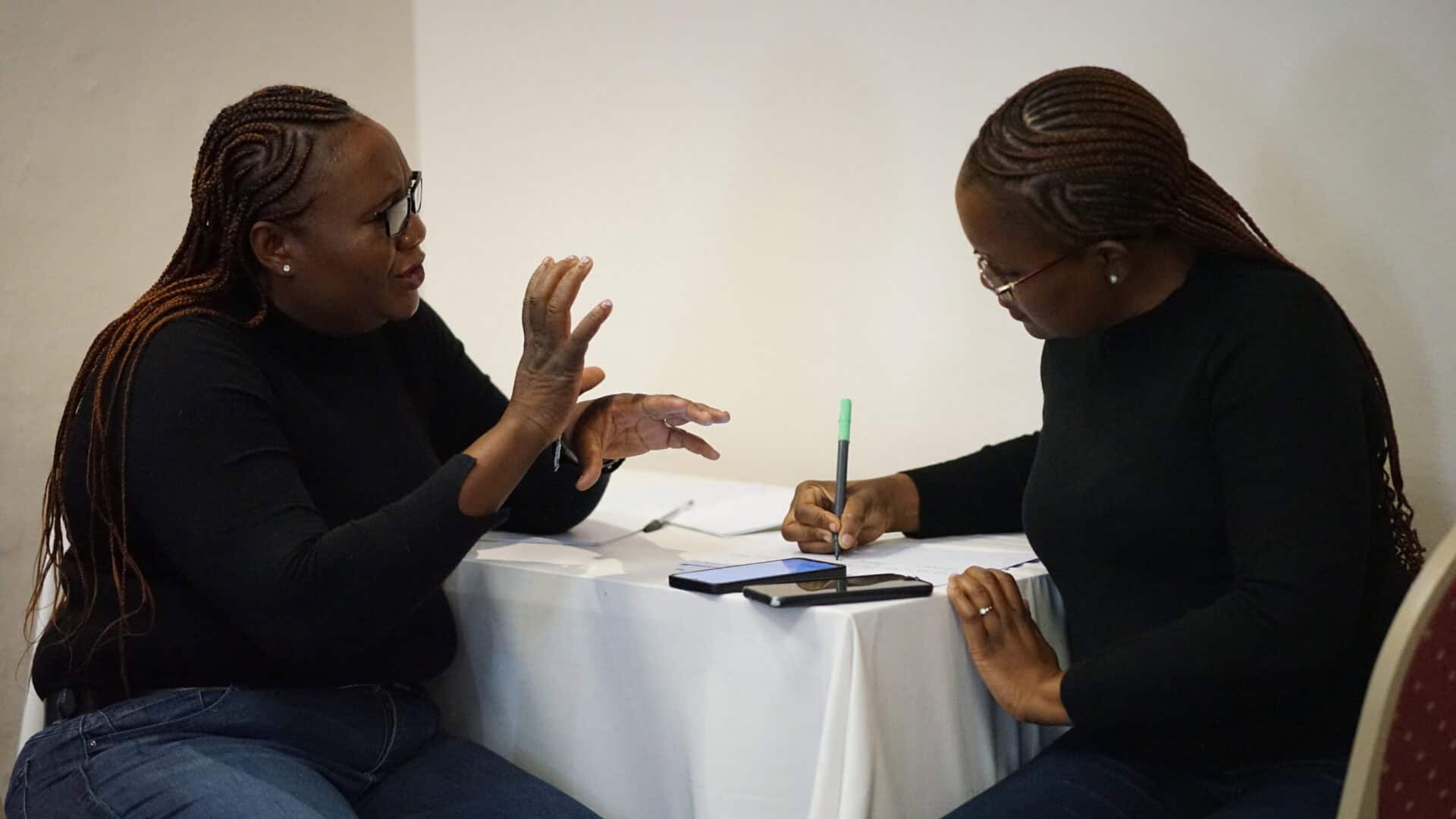 Two women sit at a table; one is speaking and gesturing with her hands, while the other is taking notes with a pen on paper.