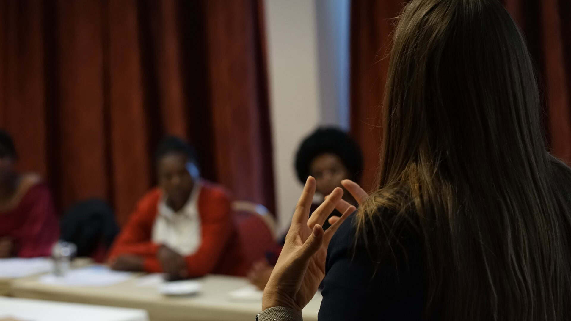 A person gestures whilst speaking to three people seated at a table in a room with red curtains.