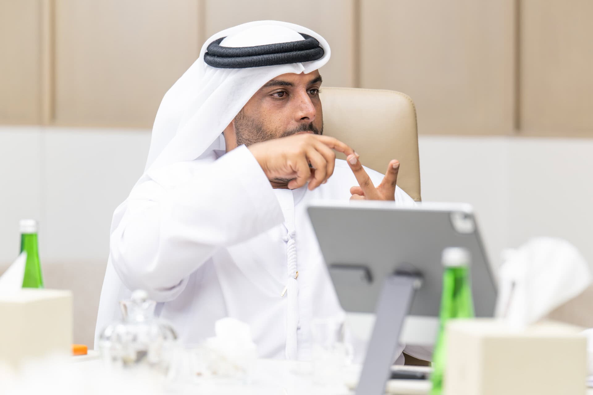 A man in traditional Middle Eastern attire sits at a table, gesturing with his hands near a tablet, with bottles and other items visible on the table.