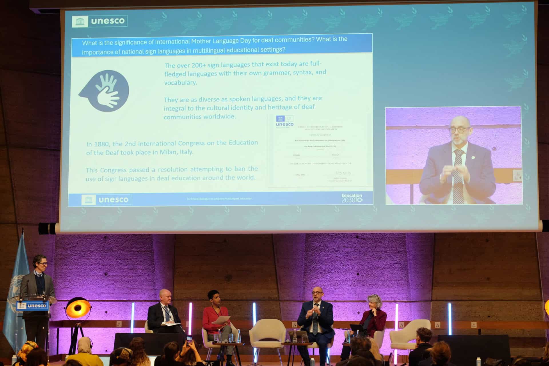 A panel of five people sits on stage discussing sign languages. A presentation slide about UNESCO and sign languages is projected behind them.