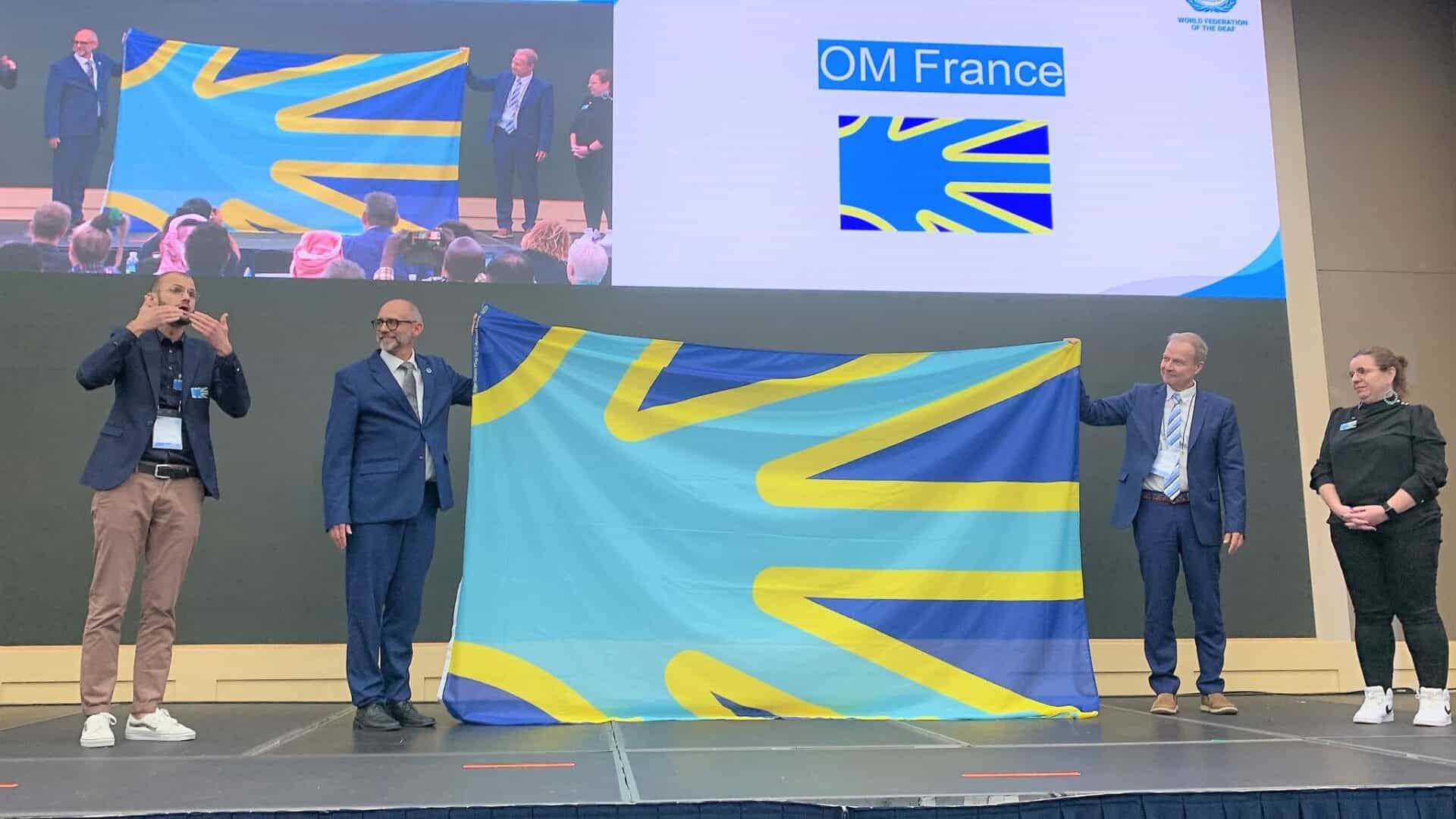 Two men hold up a large blue and yellow flag on stage while a sign language interpreter gestures to the side, with a projection screen in the background showing the flag and the text “OM France”.