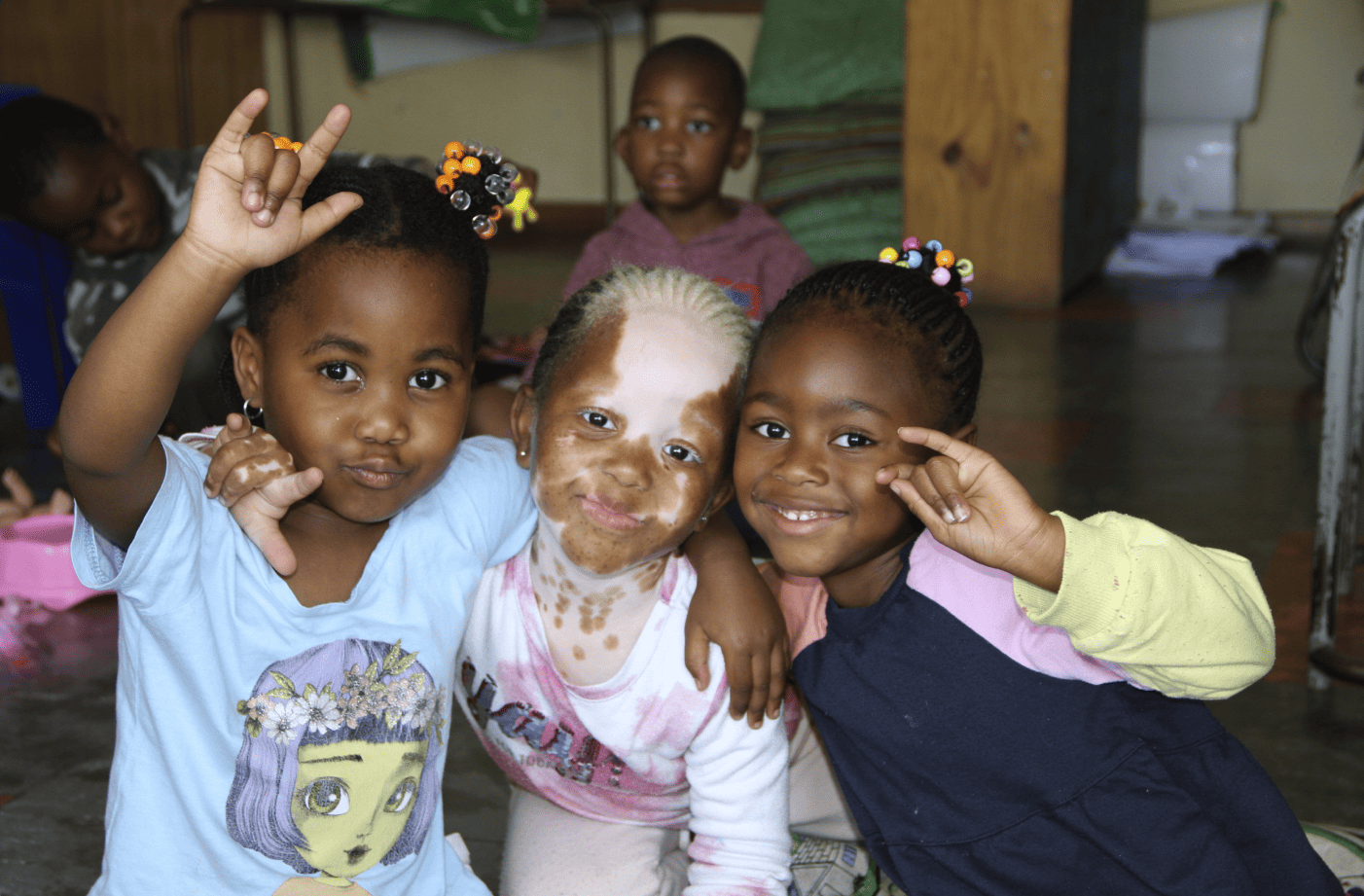 Three young children sit closely together on the floor, smiling and making hand signs, with other children and toys visible in the background.
