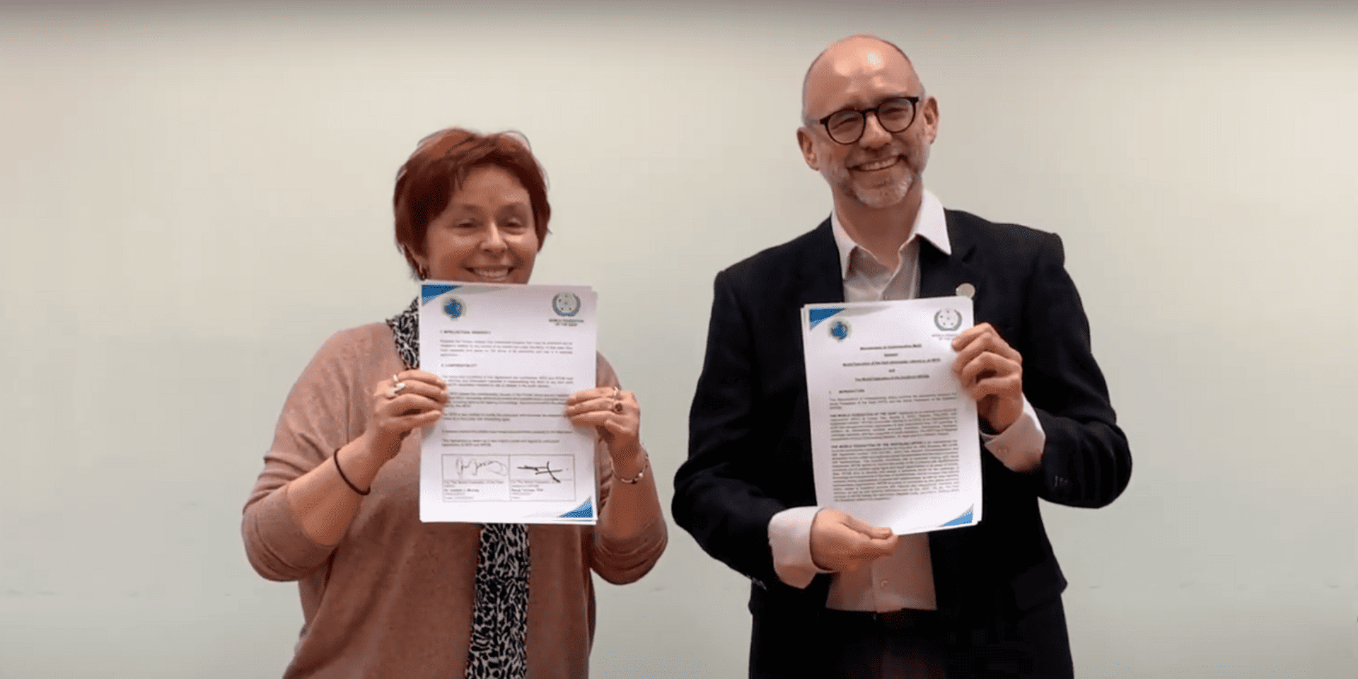 Two people smiling and holding up signed documents with official logos against a plain background.