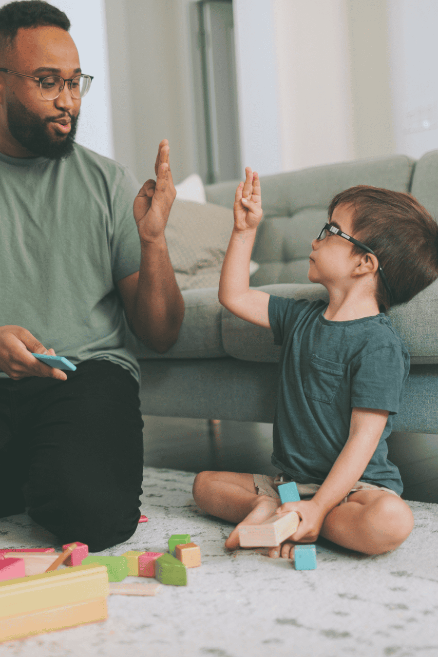 An adult and a child sit on the floor by a sofa, holding wooden blocks and giving each other a high five.