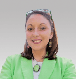 Woman in a green blazer smiles at the camera against a gray background. She has brown hair and is wearing colorful earrings and a necklace.