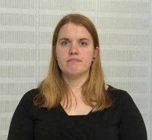 A woman with long blonde hair wearing a black top stands against a light-colored, textured wall, looking forward with a neutral expression.