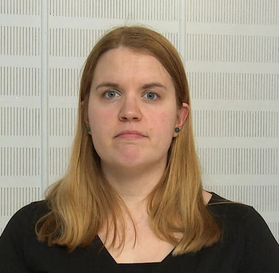 Person with long blond hair wearing a black shirt standing against a white textured wall, looking at the camera.