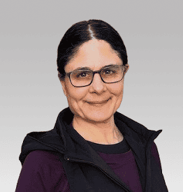 A middle-aged woman with glasses and dark hair pulled back, wearing a dark vest over a maroon top, stands against a gray gradient background.