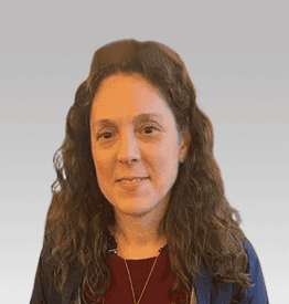 A person with long, curly brown hair is smiling slightly, wearing a dark top against a plain background.