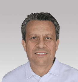 A man with short, dark hair and a light complexion wearing a white shirt smiles slightly against a gray background.