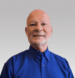 Bald man with a trimmed white beard wearing a blue button-up shirt, standing in front of a plain light gray background.