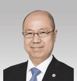 A person wearing glasses, a dark striped suit, and a blue tie is smiling against a light gray background.