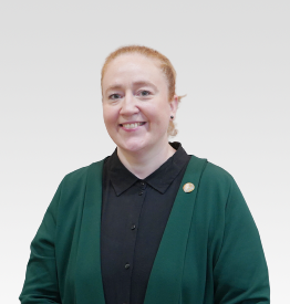 A person wearing a green cardigan over a black shirt stands against a light gray background, smiling.
