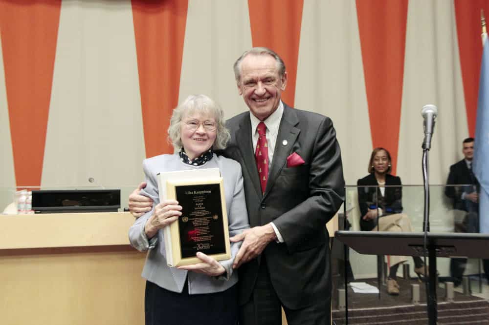 Two people stand together in a room with orange curtains. One person holds a framed plaque. Others are seated in the background.