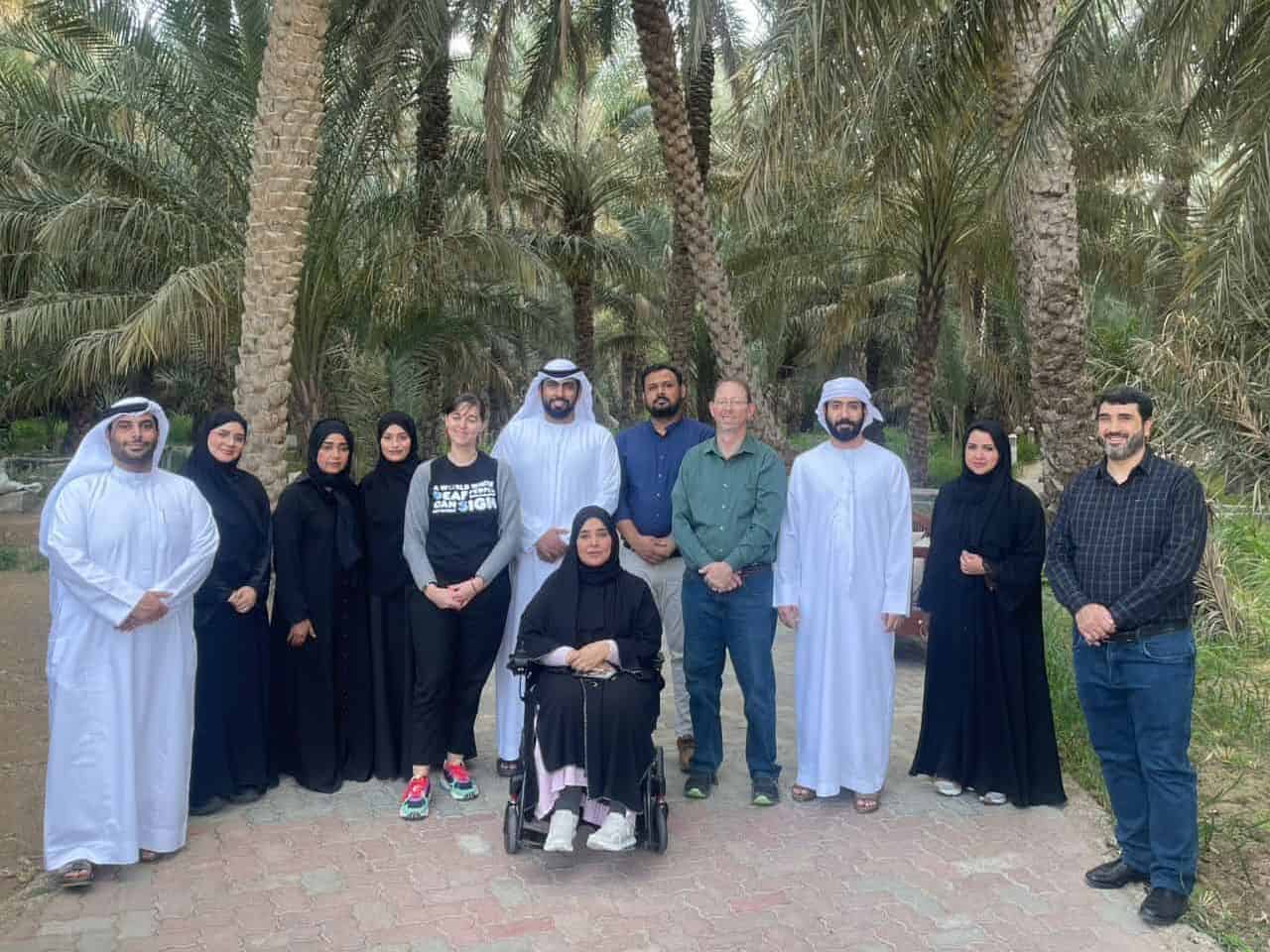 A group of eleven people, including one person in a wheelchair, pose together on a paved path in a palm tree grove.