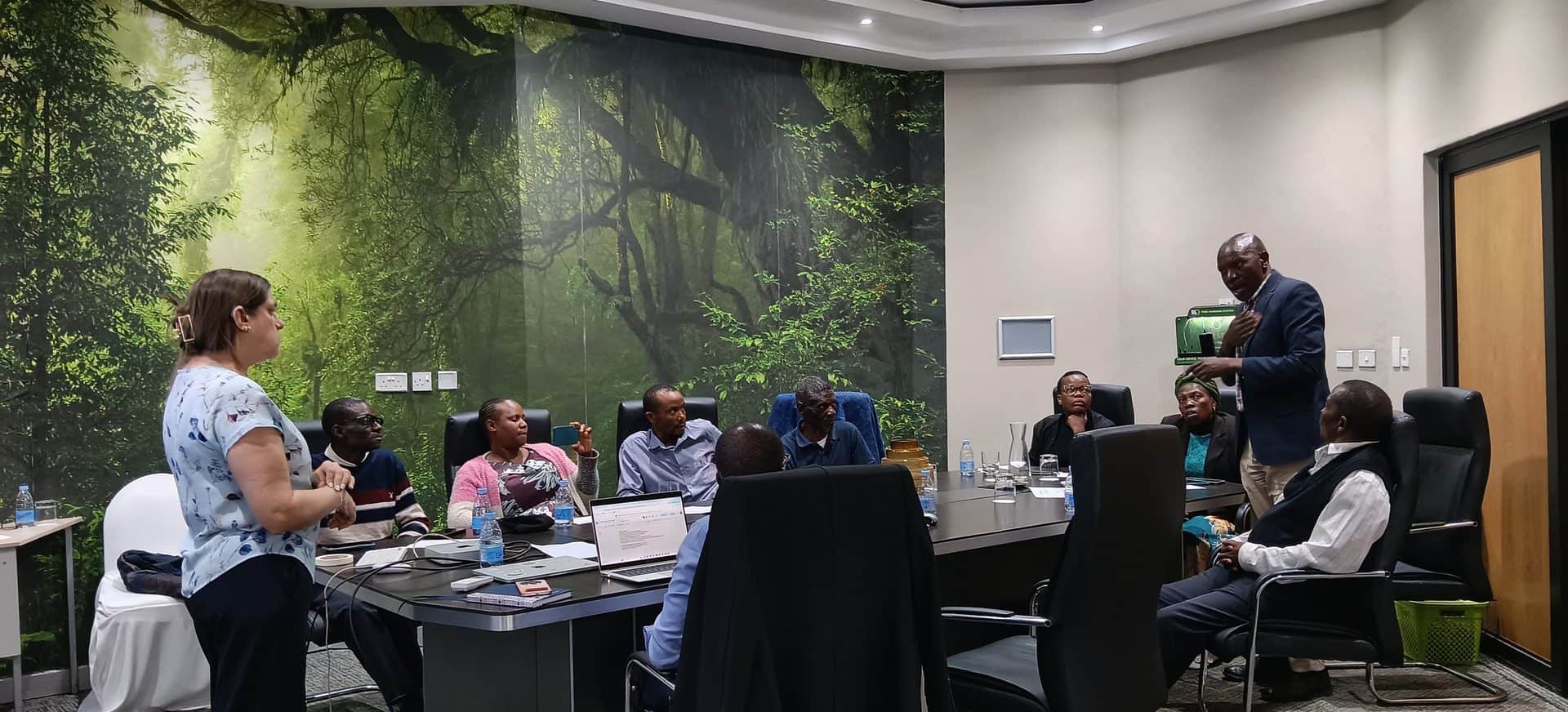 A woman stands presenting to a group of people seated around a conference table in a room featuring a large mural of a forest.