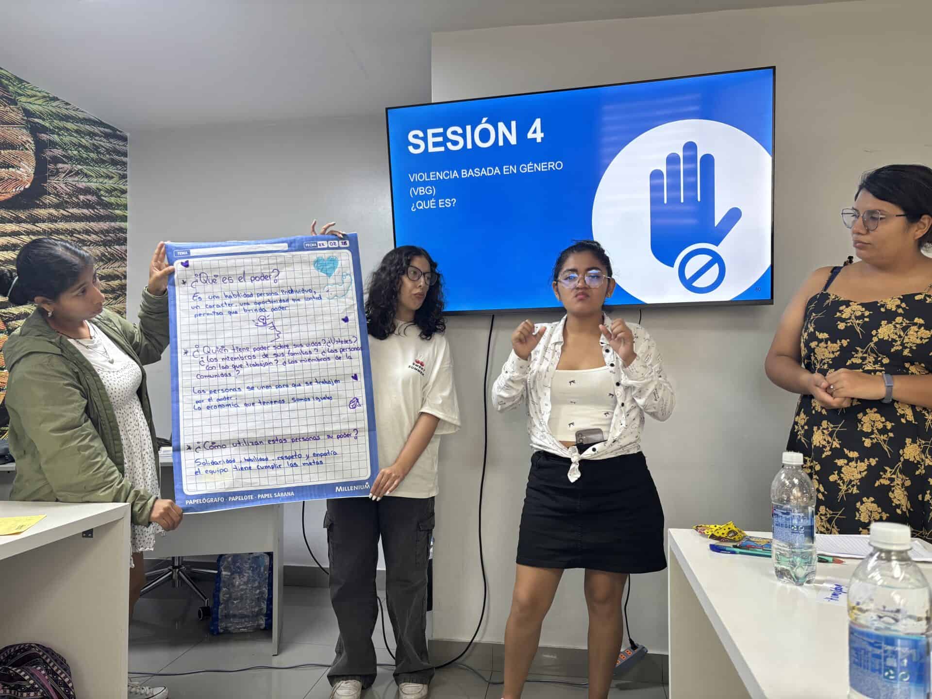 Four women stand in front of a screen displaying “Sesión 4: Violencia Basada en Género (VBG)” while one speaks and another holds a large handwritten poster.