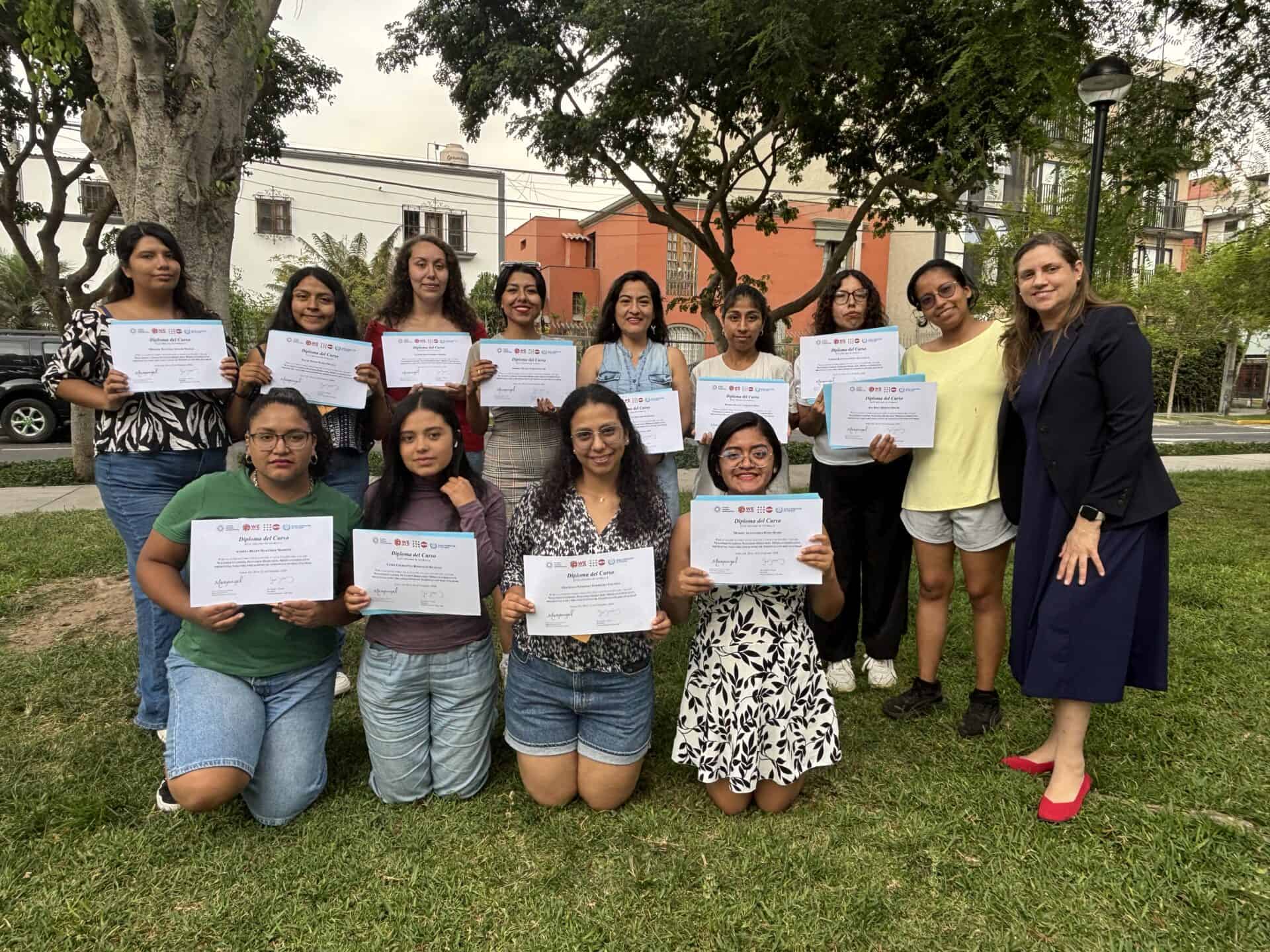 A group of women pose outdoors, holding certificates, with one woman standing to the side. Trees, grass, and buildings are visible in the background.
