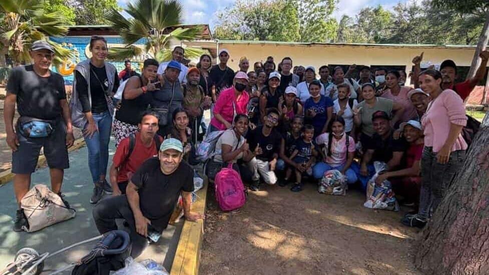 A large group of people gathers outdoors near trees and a yellow building, many smiling at the camera. Some are holding bags and standing or sitting on a paved area.