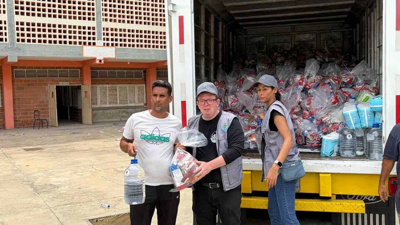Three people distribute bags of supplies and bottled water from a lorry loaded with relief packages in an outdoor area.
