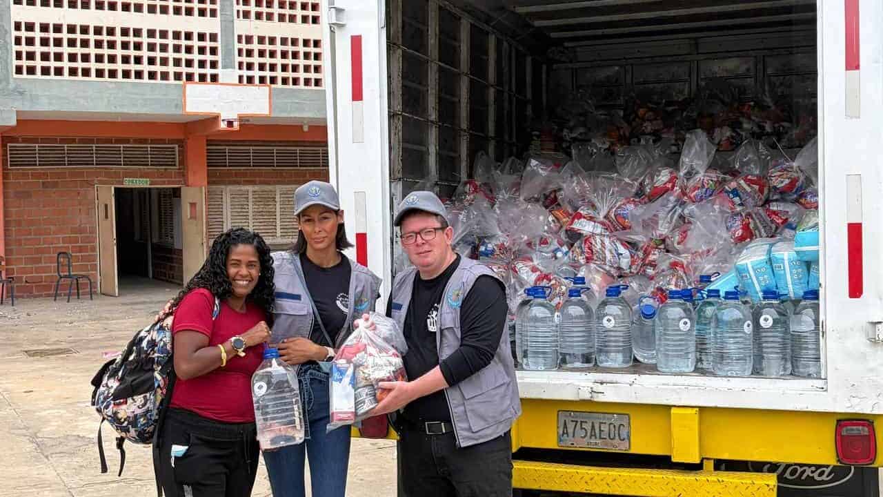 Three people stand in front of an open lorry filled with bags containing bottled water and food supplies, holding some of the bags and smiling at the camera.