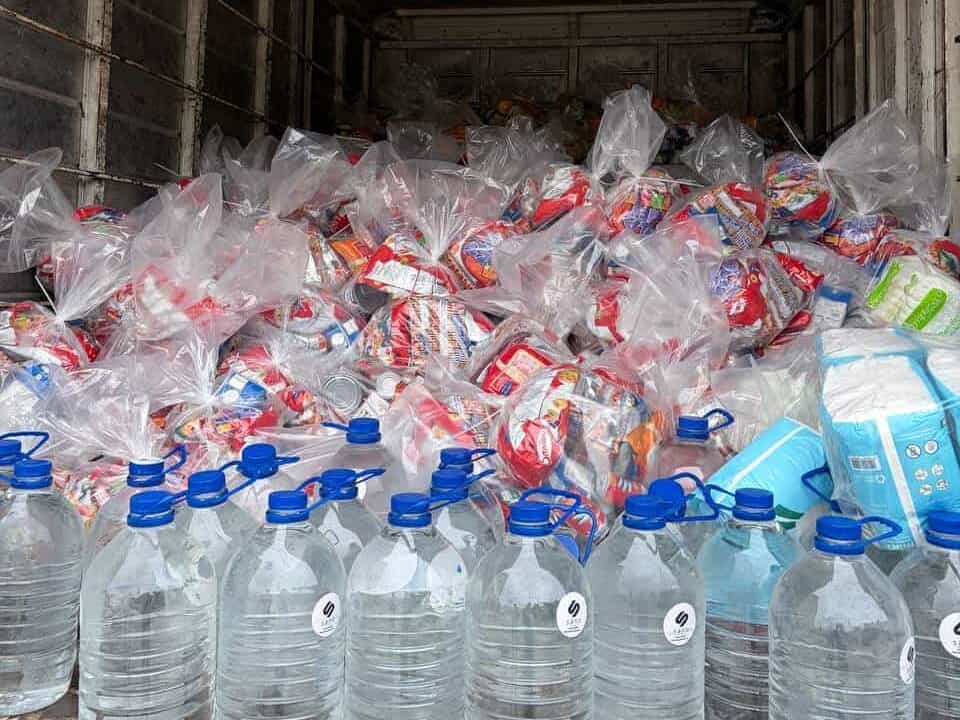 Large water bottles and plastic bags filled with food items are arranged in the back of a lorry.
