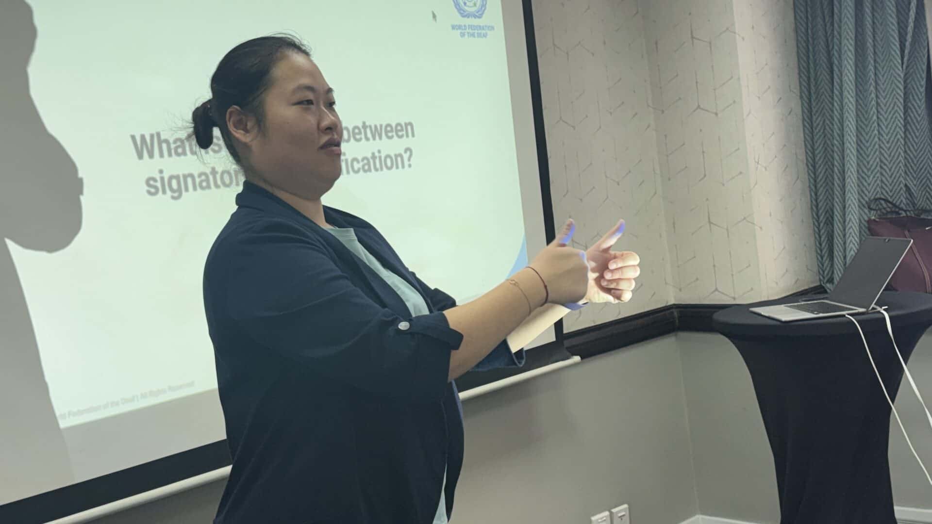 A woman is using sign language in front of a projected presentation slide in a conference room.