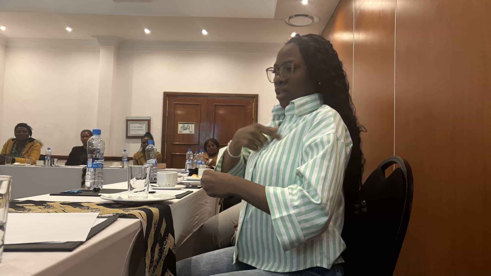 A woman in a green striped shirt speaks at a conference table, whilst others sit and listen in a meeting room with wooden walls and bright lighting.
