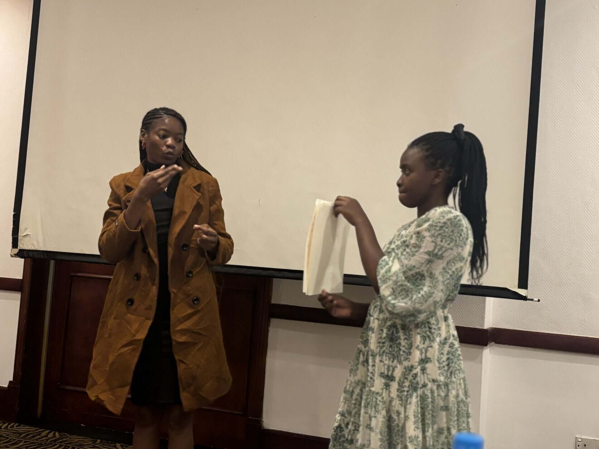 Two women stand in front of a projector screen; one gestures whilst speaking, and the other holds a white cloth or paper.