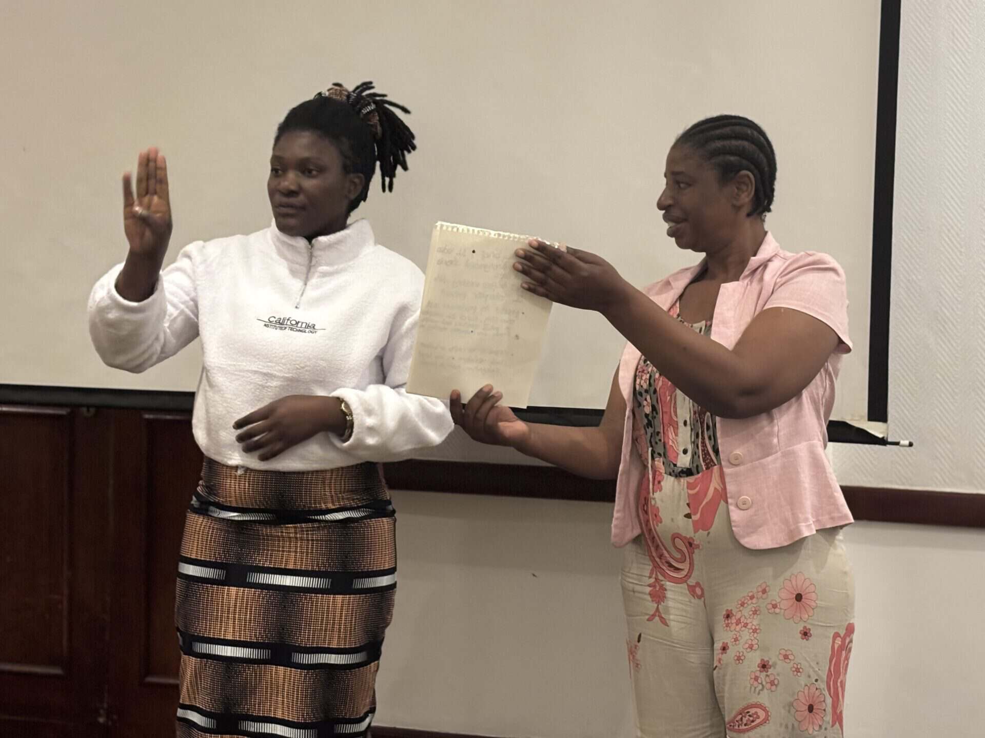 Two women stand indoors; one raises her right hand, while the other holds a handwritten paper. Both face forwards, with a blank projector screen in the background.