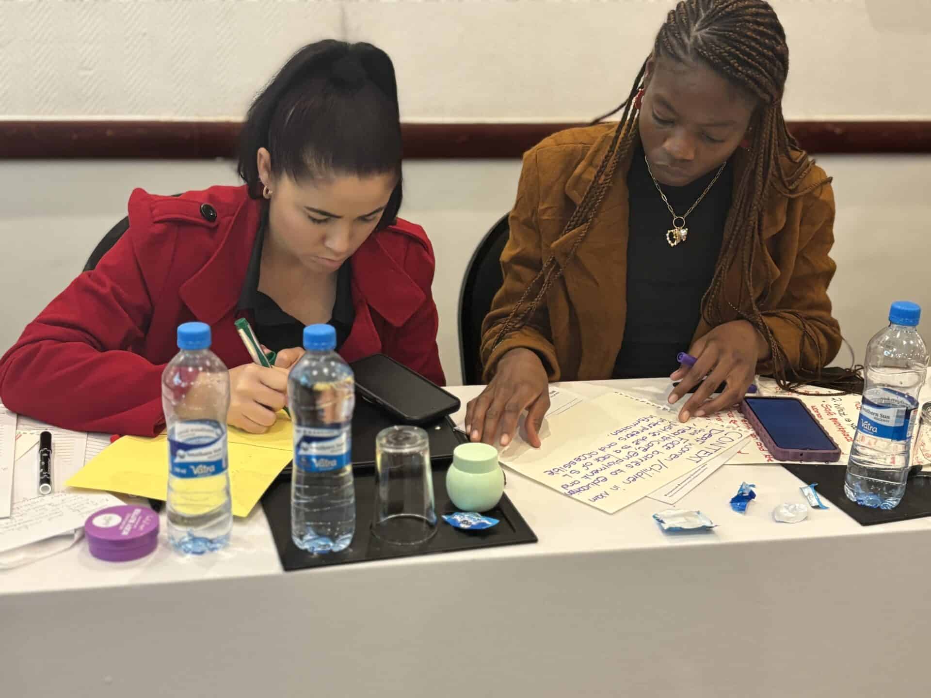 Two women sit at a table with notebooks, writing and working together. Bottled water, snacks, and papers are spread out on the table in front of them.