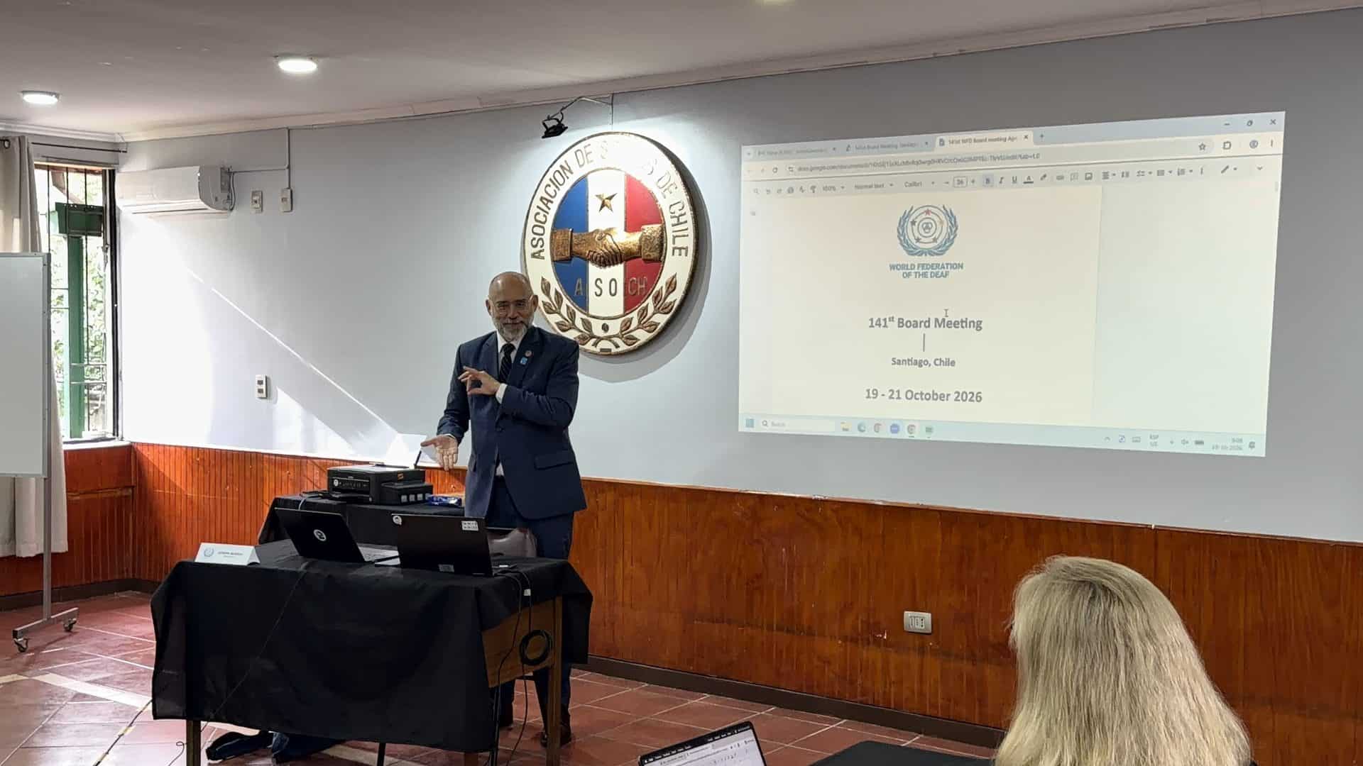 A man in a suit signs whilst presenting in front of a projection screen that displays text for a World Federation of the Deaf board meeting in Santiago, Chile.