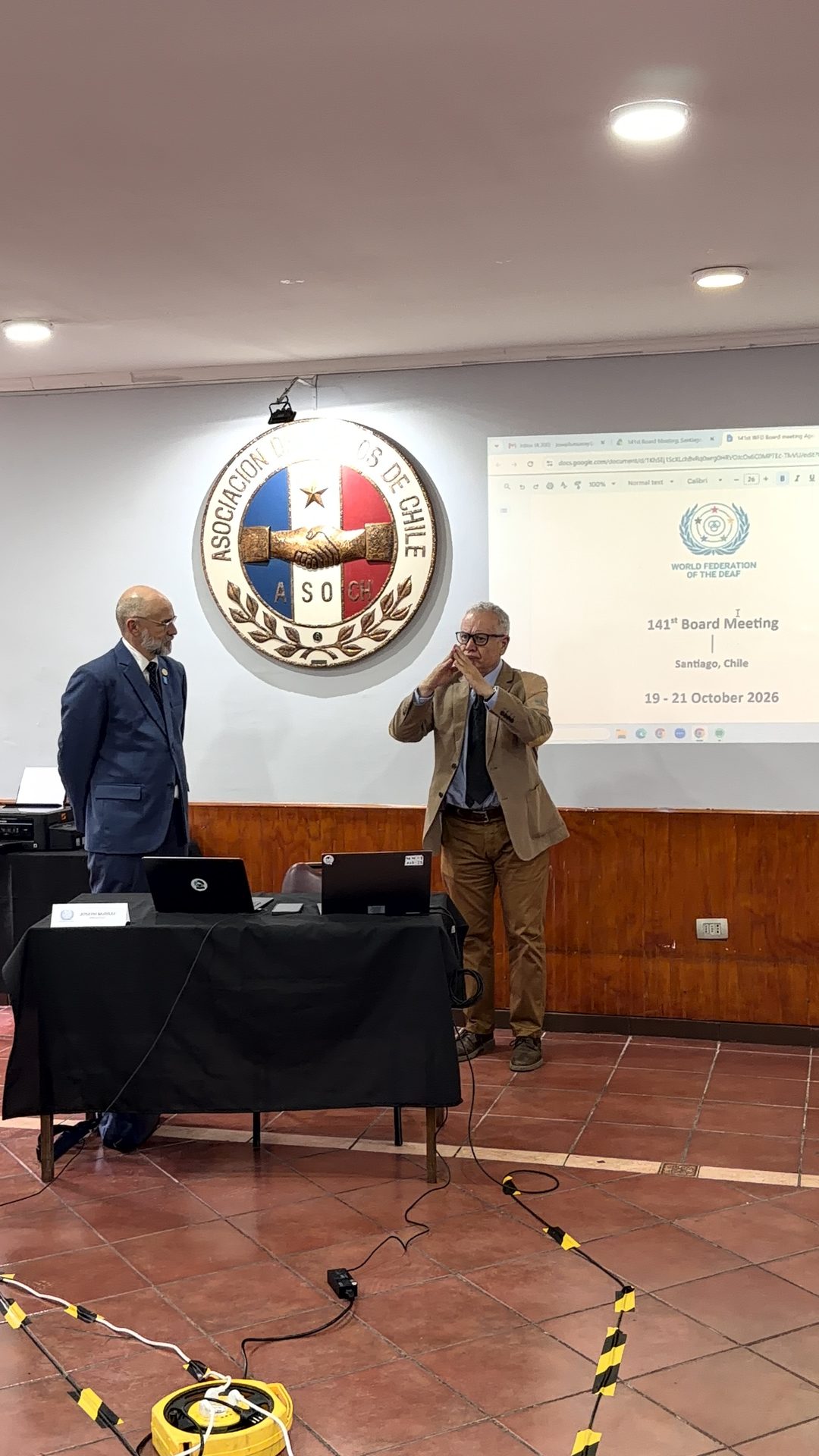 Two men stand in front of a projection screen displaying “141st Board Meeting, World Federation of the Deaf, Santiago, Chile, 19–21 October 2026.” One man is signing.