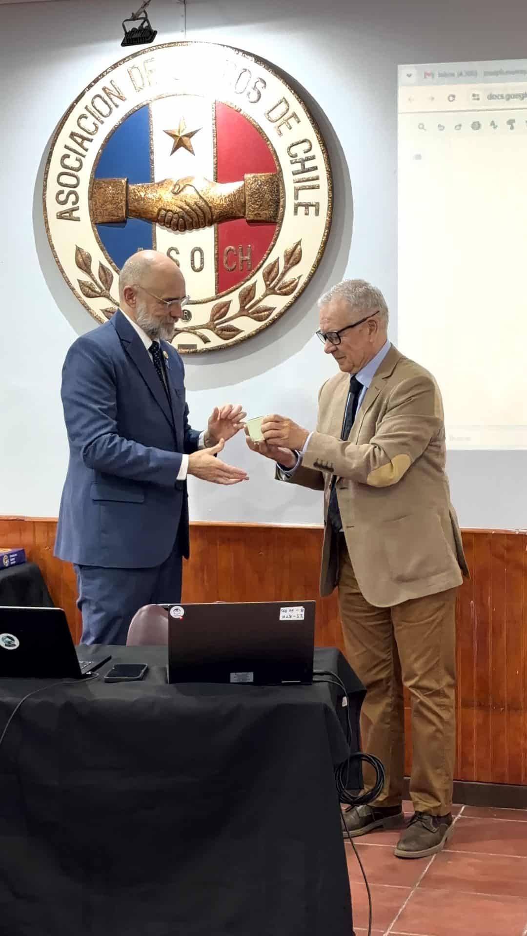 Two men in blazers standing at a table, one handing a small rectangular object to the other in front of the Asociación de Empleados de Chile emblem.