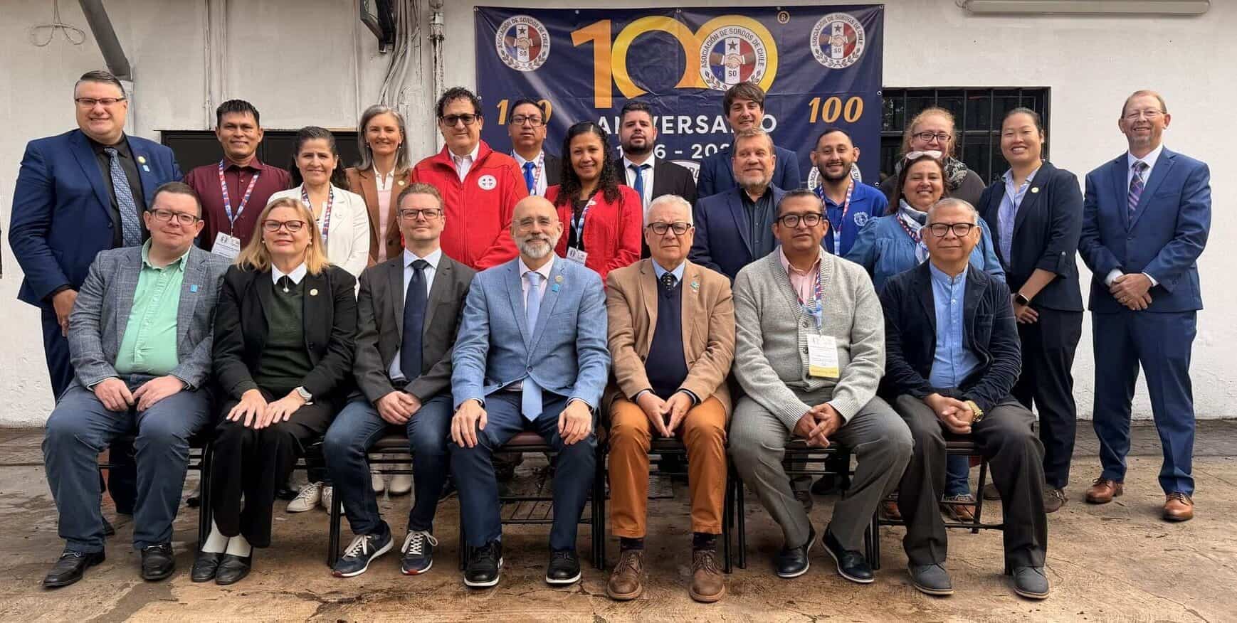 A group of people posing for a photo in front of a banner celebrating the 100th anniversary of the Asociación de Sordos de Chile.