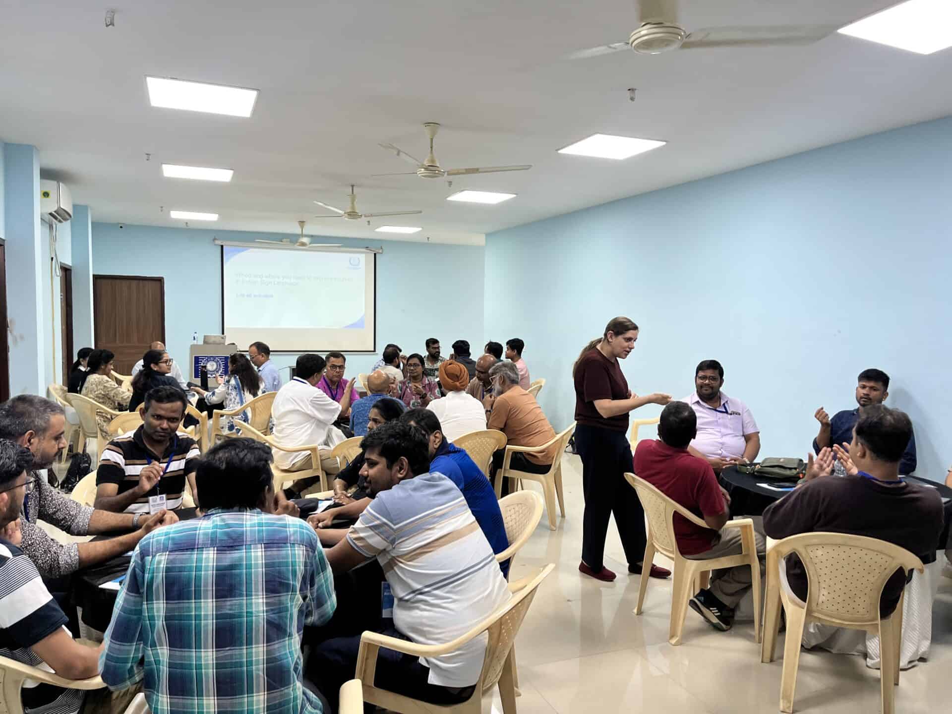 A group of people are seated at round tables in a bright conference room, engaging in discussions whilst a presentation is displayed on a projector screen.
