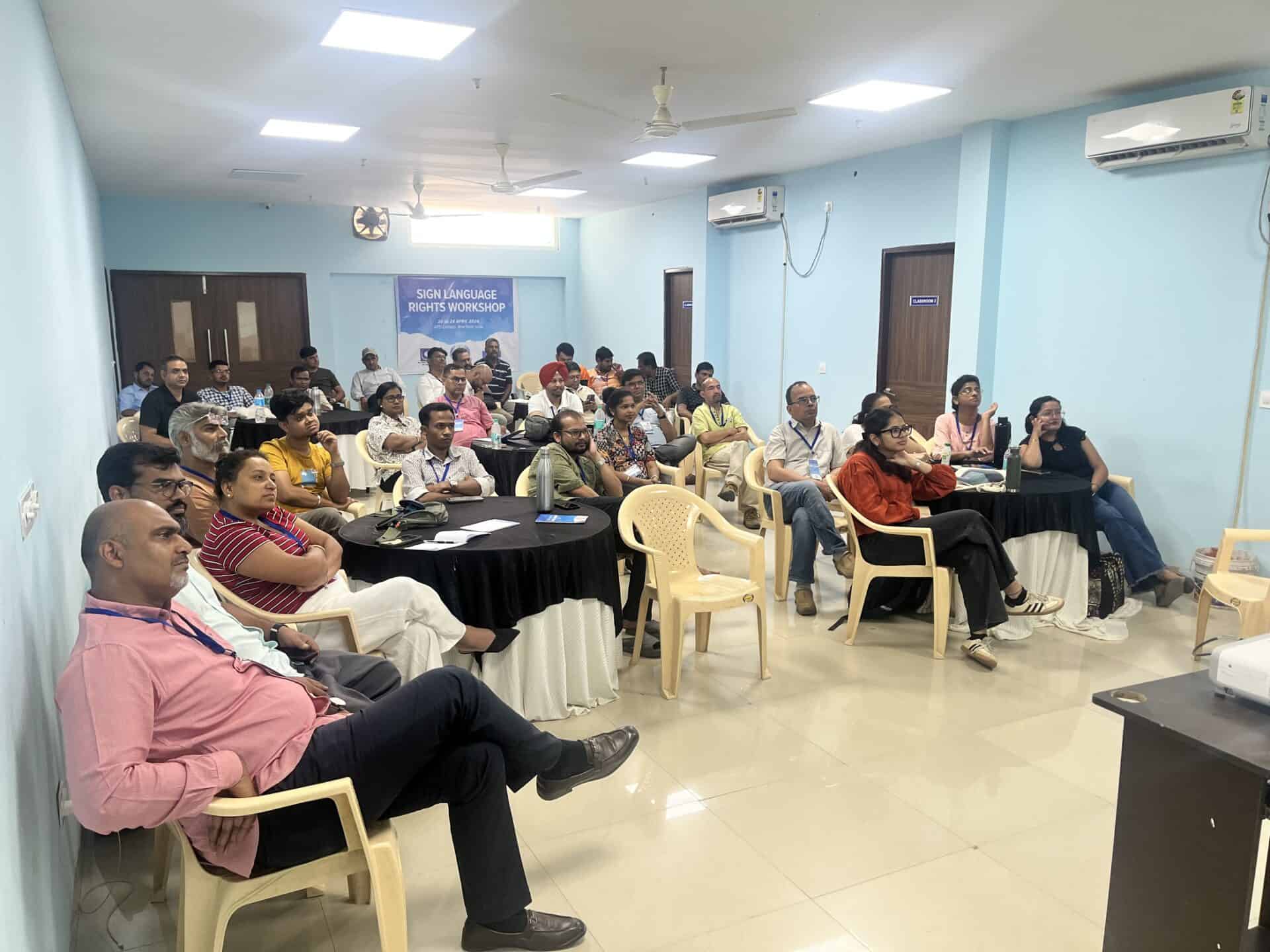 A group of adults sits in a classroom, listening to a presentation. Tables are arranged in clusters, and a sign reads "Sign Language Rights Workshop.