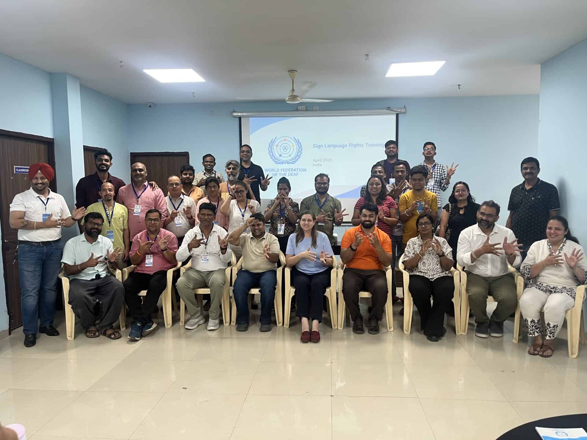 A group of people pose indoors, some seated and some standing, in front of a screen displaying "Sign Language Rights Training." Several individuals hold up their hands in a gesture.