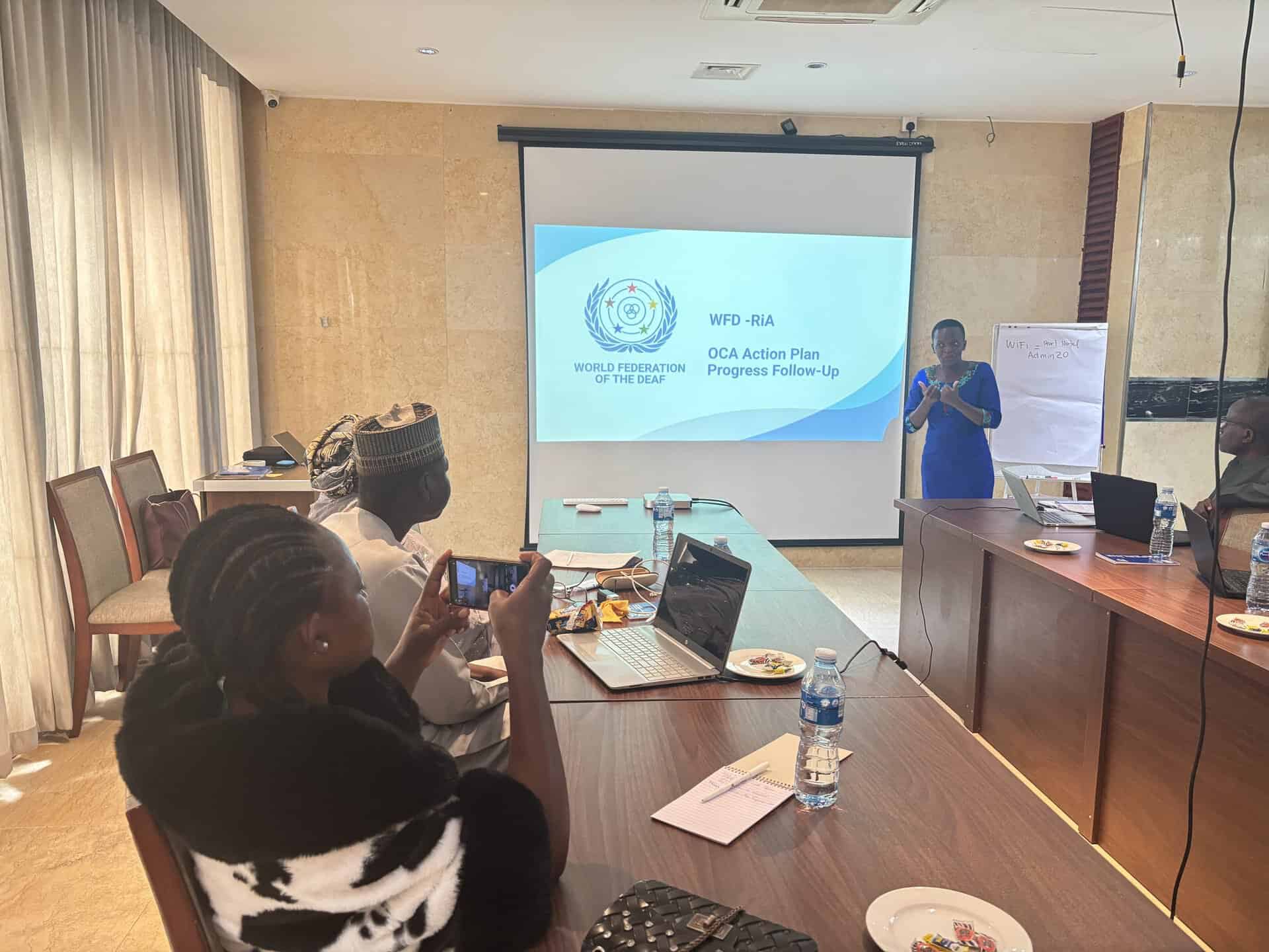 A woman in a blue dress presenting to a group in a conference room, with a screen showing the World Federation of the Deaf logo and text about the OCA Action Plan.