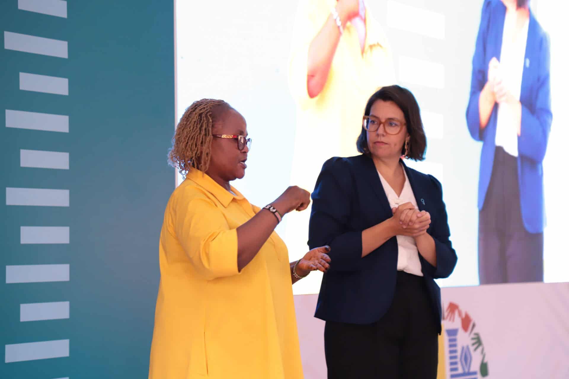Two women stand on stage, one wearing a yellow shirt and speaking while gesturing, and the other in a dark blazer watching attentively. A large screen displays one of the women in the background.