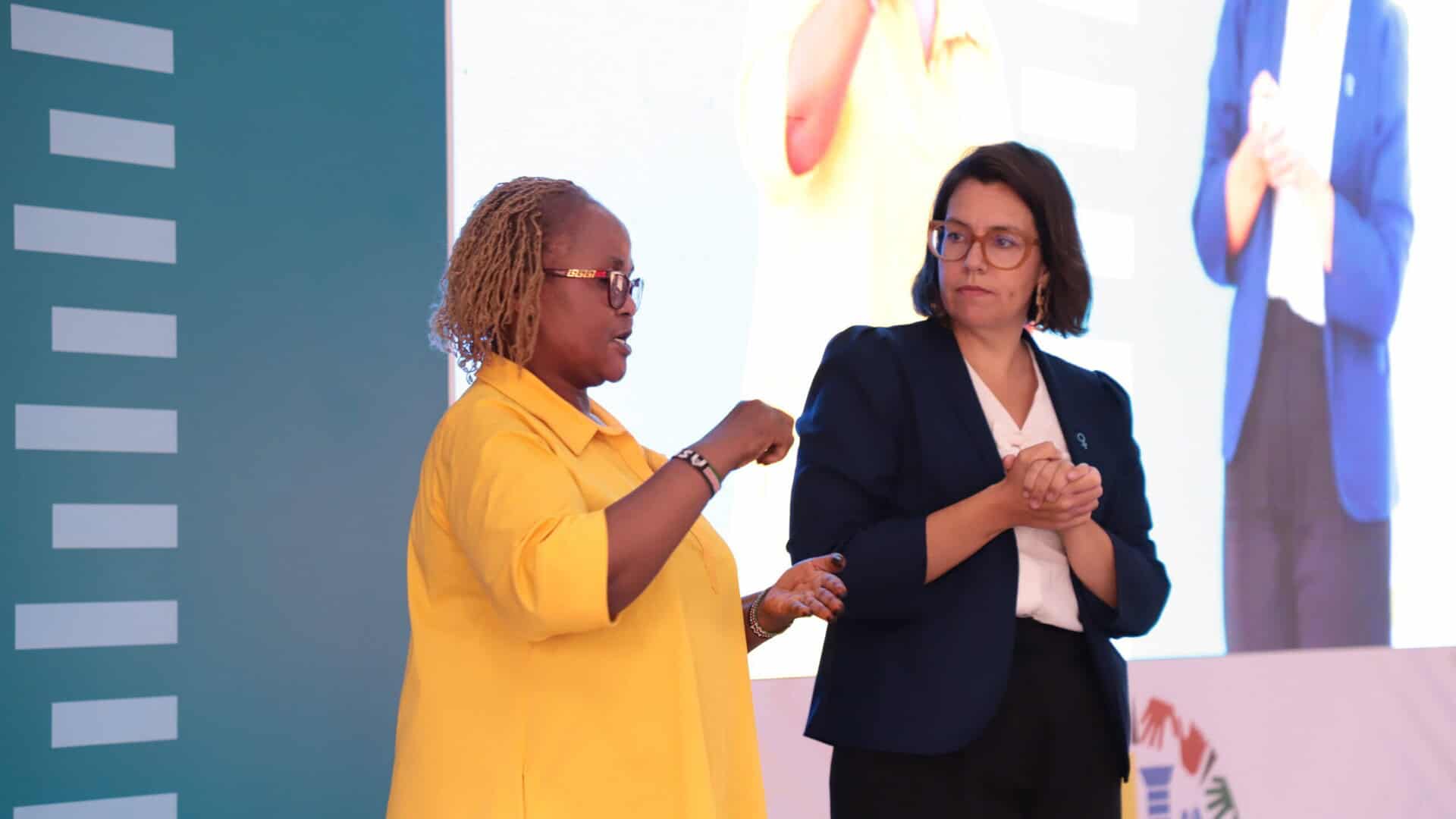Two women stand on stage, one wearing a yellow shirt and speaking while gesturing, and the other in a dark blazer watching attentively. A large screen displays one of the women in the background.