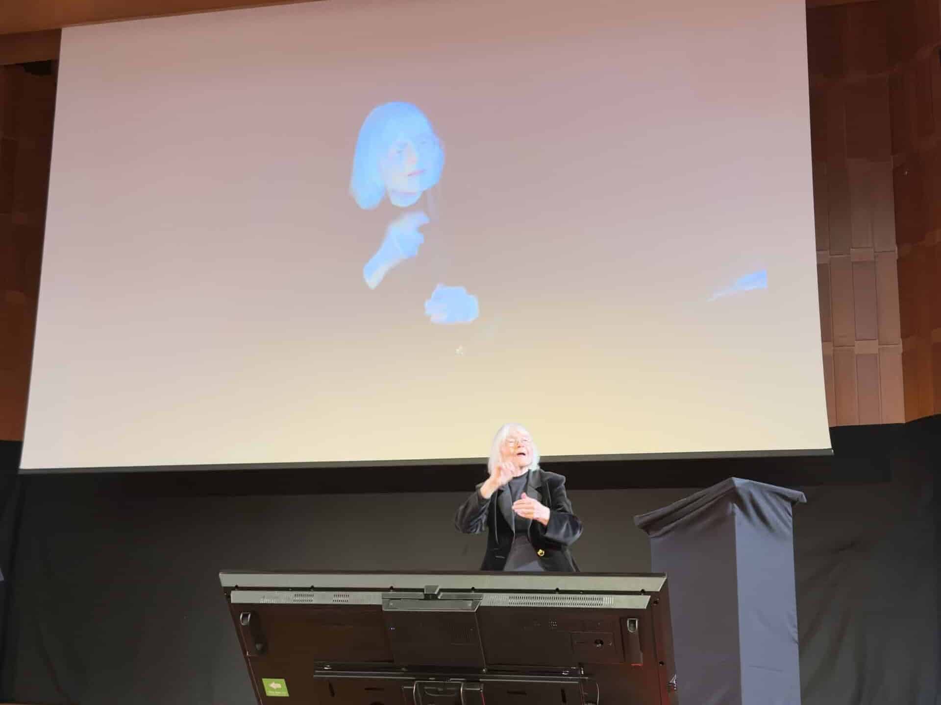 A person stands on stage signing, with their image projected on a large screen behind them in an auditorium setting.