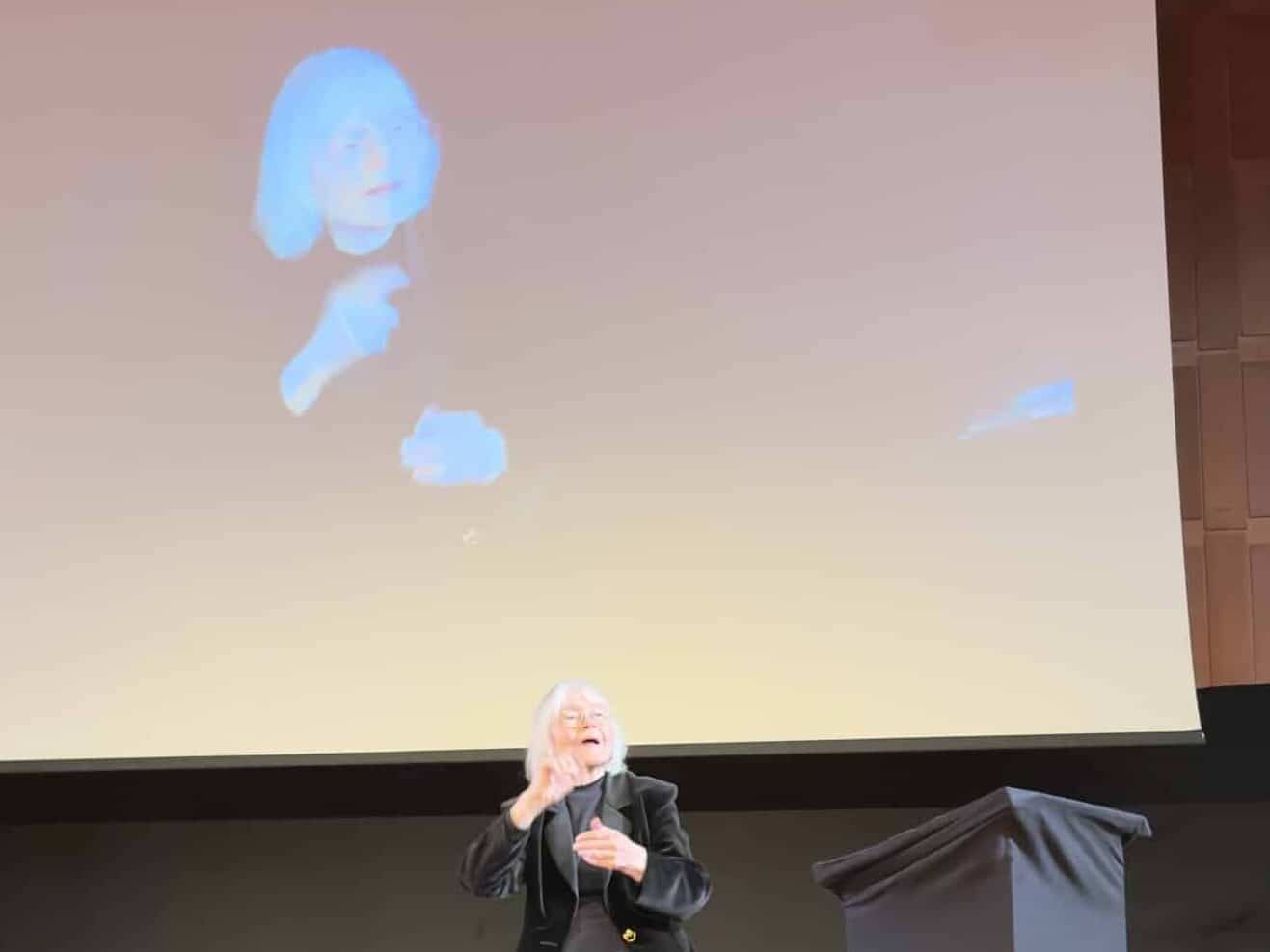 A person stands on stage signing, with their image projected on a large screen behind them in an auditorium setting.
