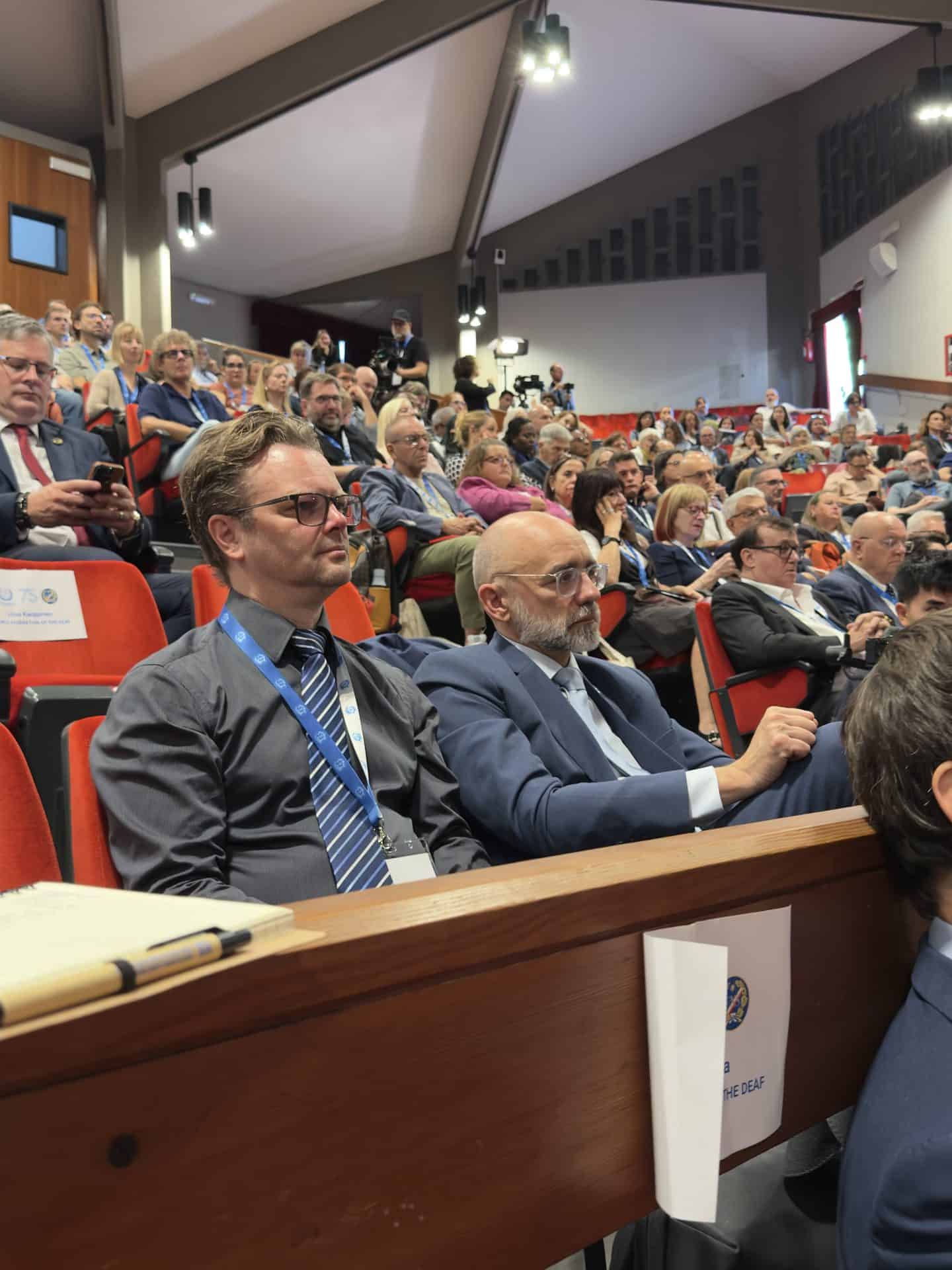Audience members, including two men in suits in the foreground, sit attentively in a large auditorium during a conference or formal event.