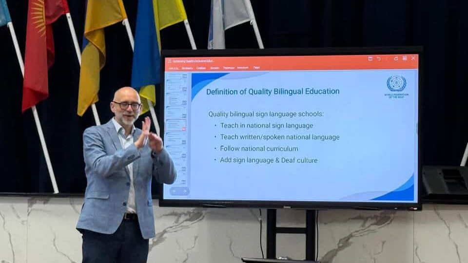 A man stands next to a screen displaying a slide titled "Definition of Quality Bilingual Education" with flags in the background.