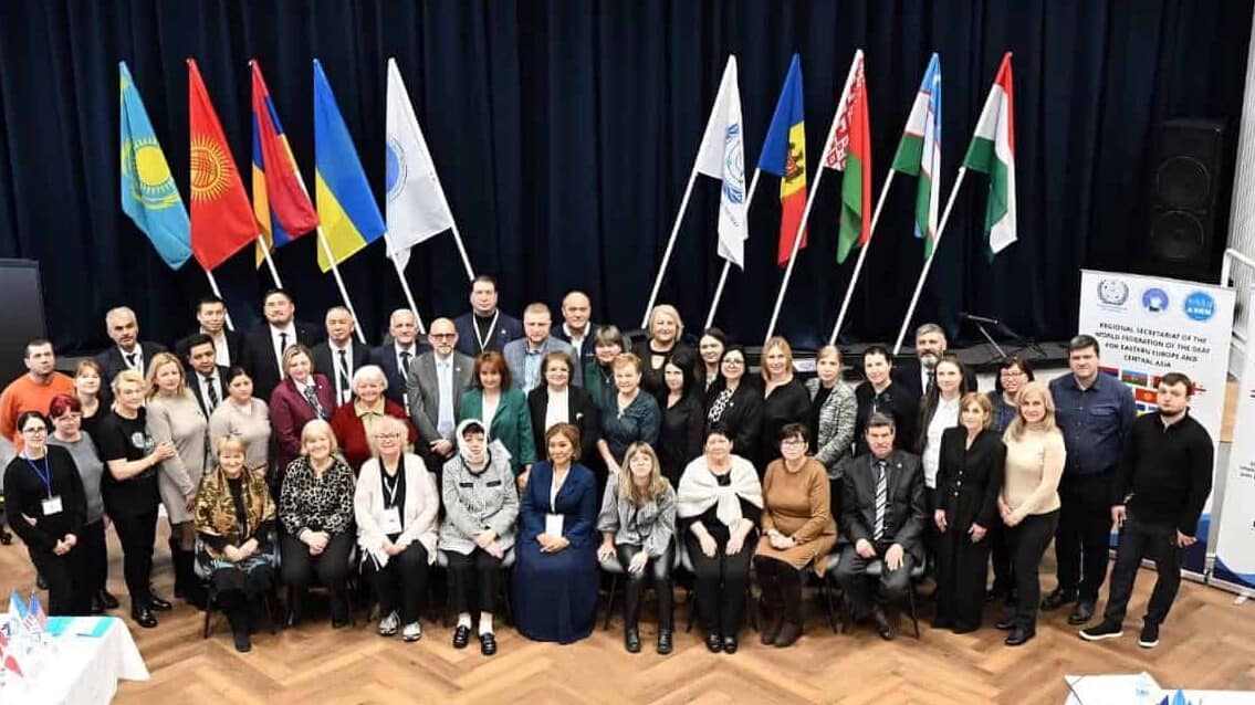 A large group of people pose for a photo in a conference room with tables, banners, and multiple national flags displayed in the background.