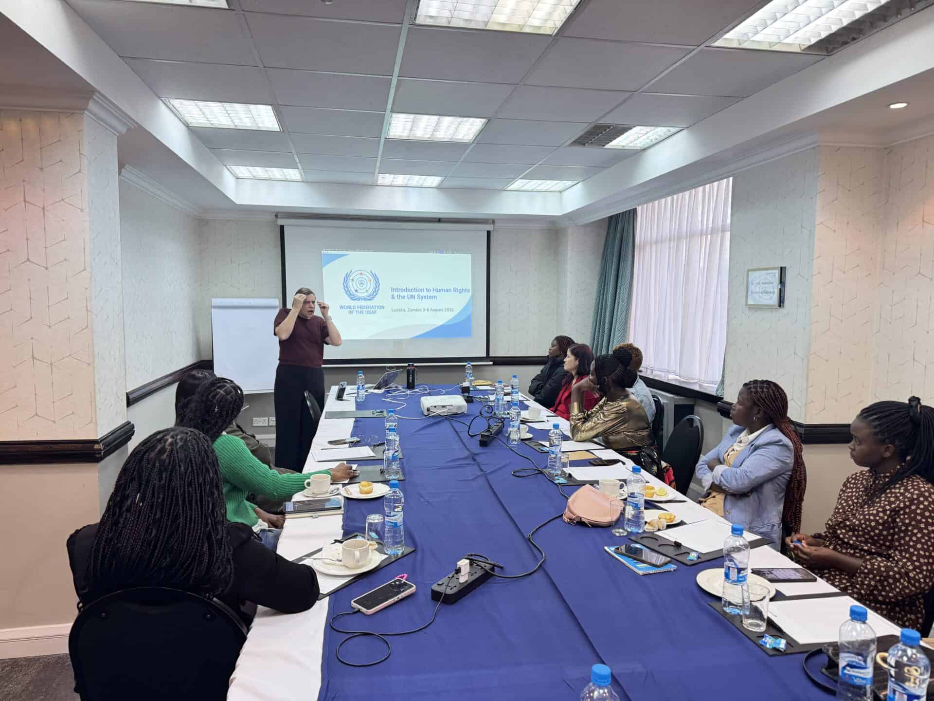 A person gives a presentation to a group of people seated around a conference table, with a slide on human rights displayed on the screen at the front of the room.