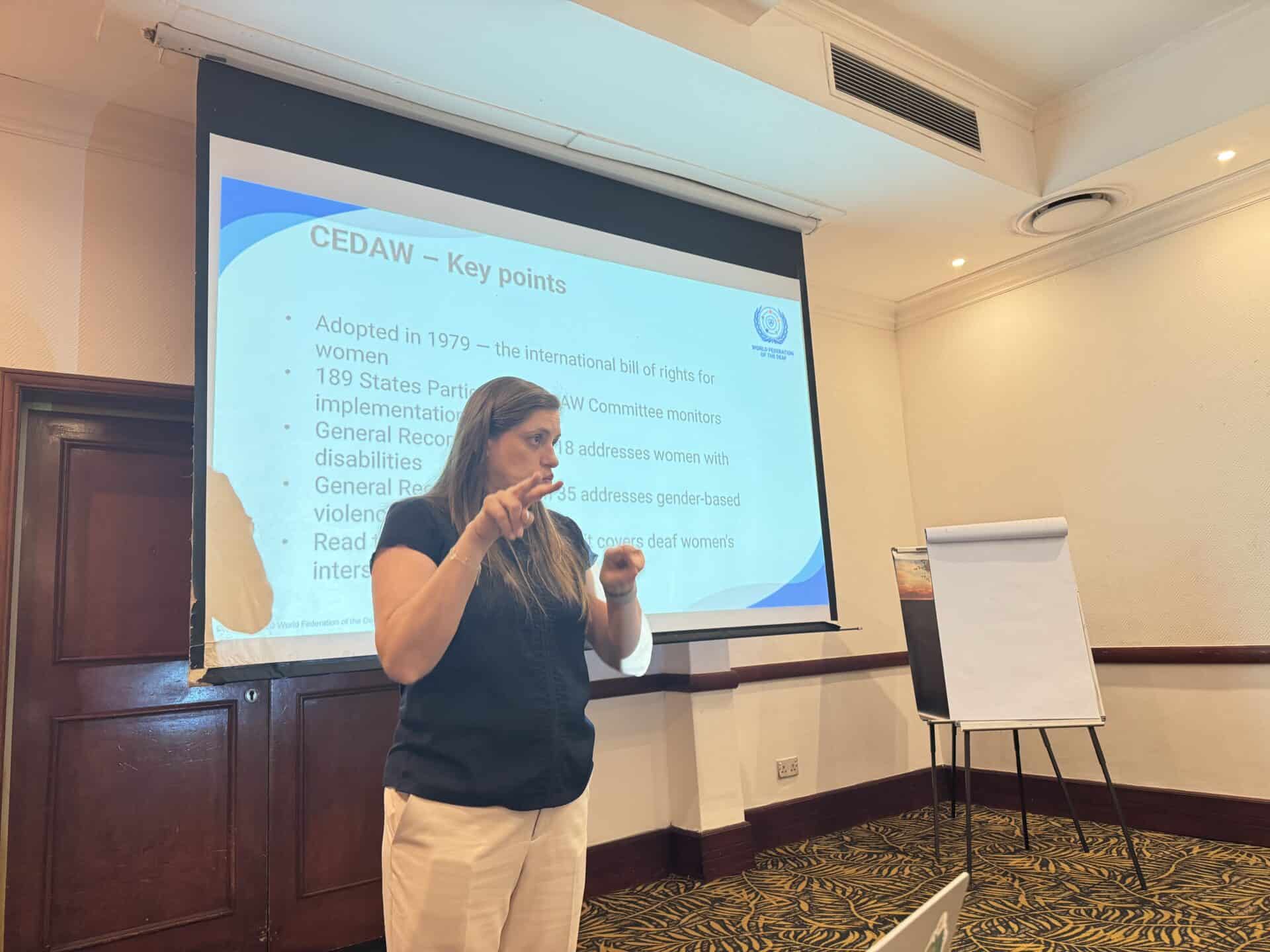 A woman stands in front of a projection screen giving a presentation on CEDAW. The screen displays key points, and an empty flipchart is visible to the side.