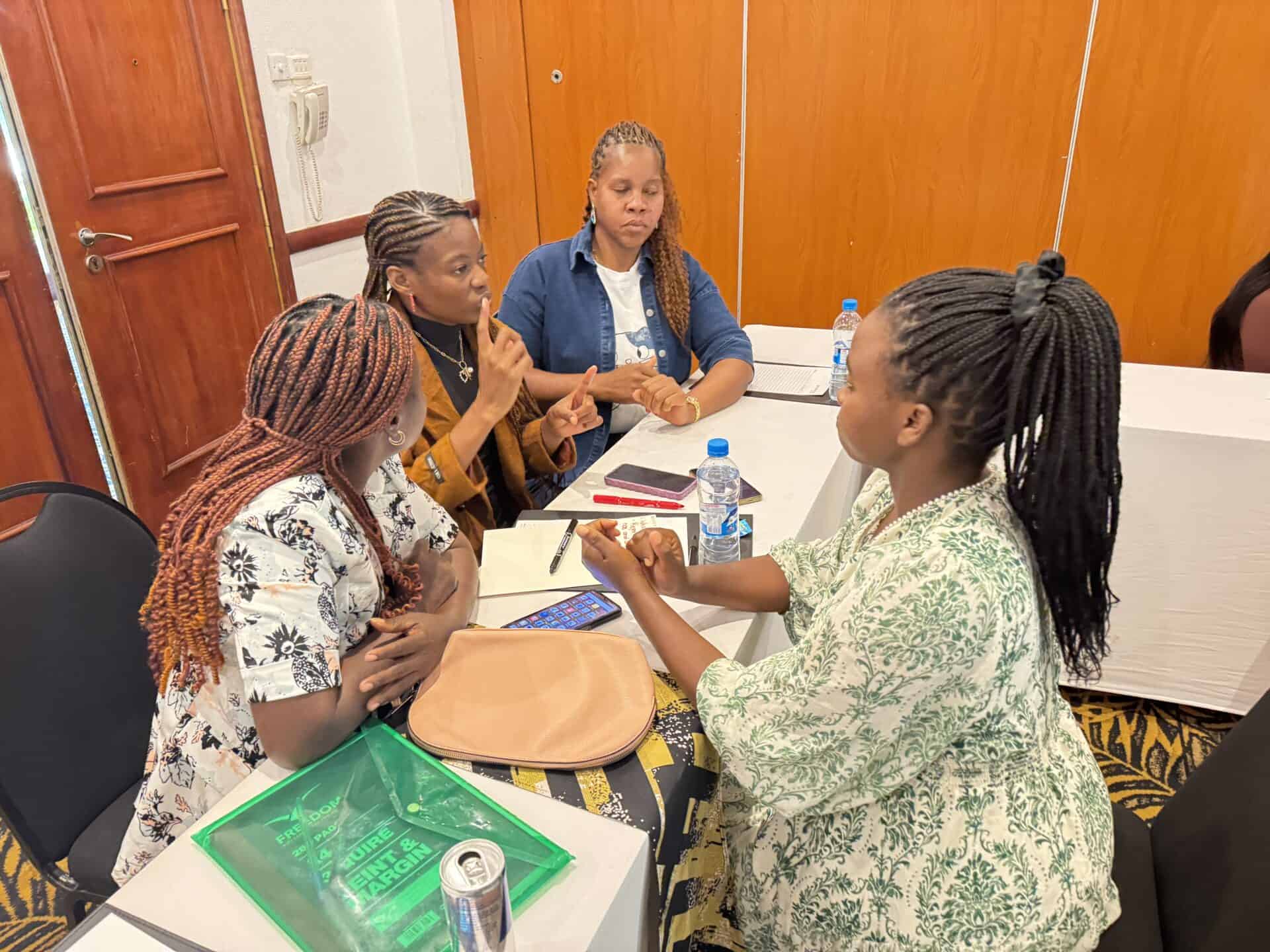 Four women sit around a table engaged in discussion, with notebooks, a mobile phone, a drinks can, and bottled water on the table in a meeting room.