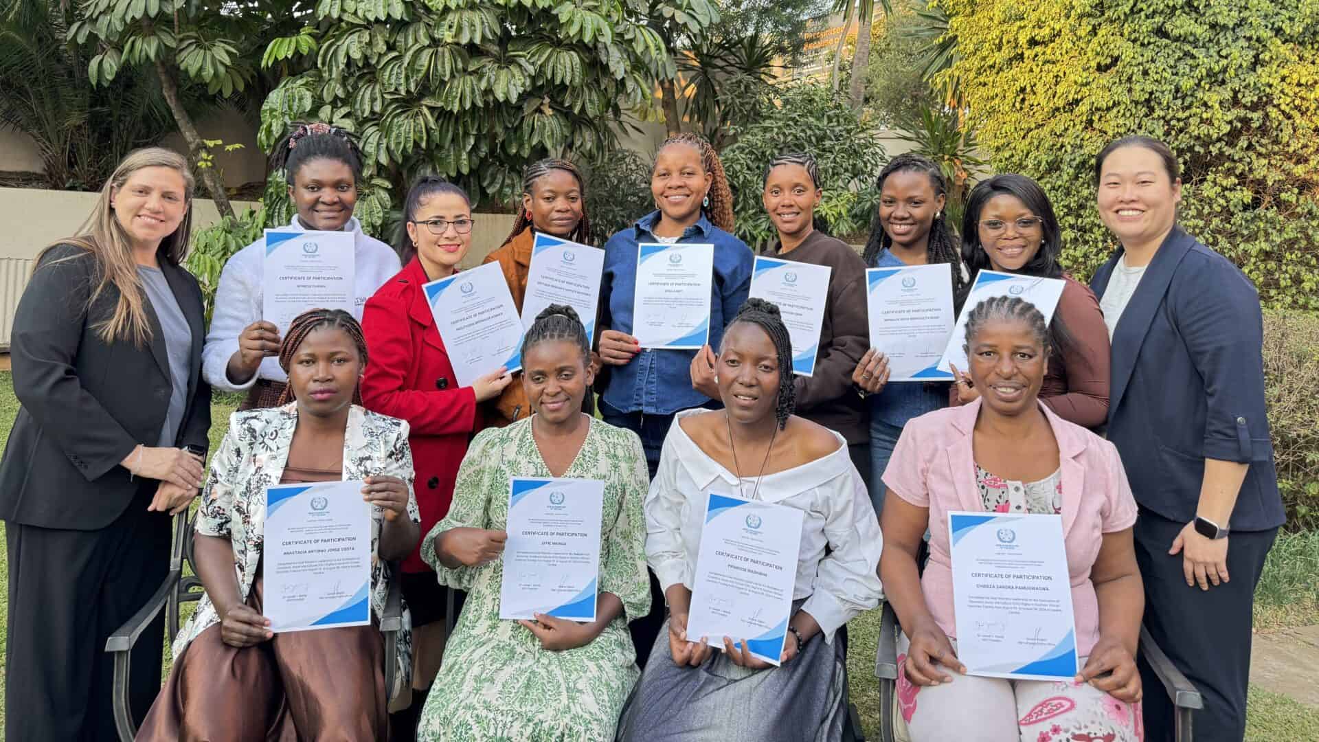 A group of twelve women pose outdoors, holding certificates and smiling at the camera, with greenery and trees in the background.